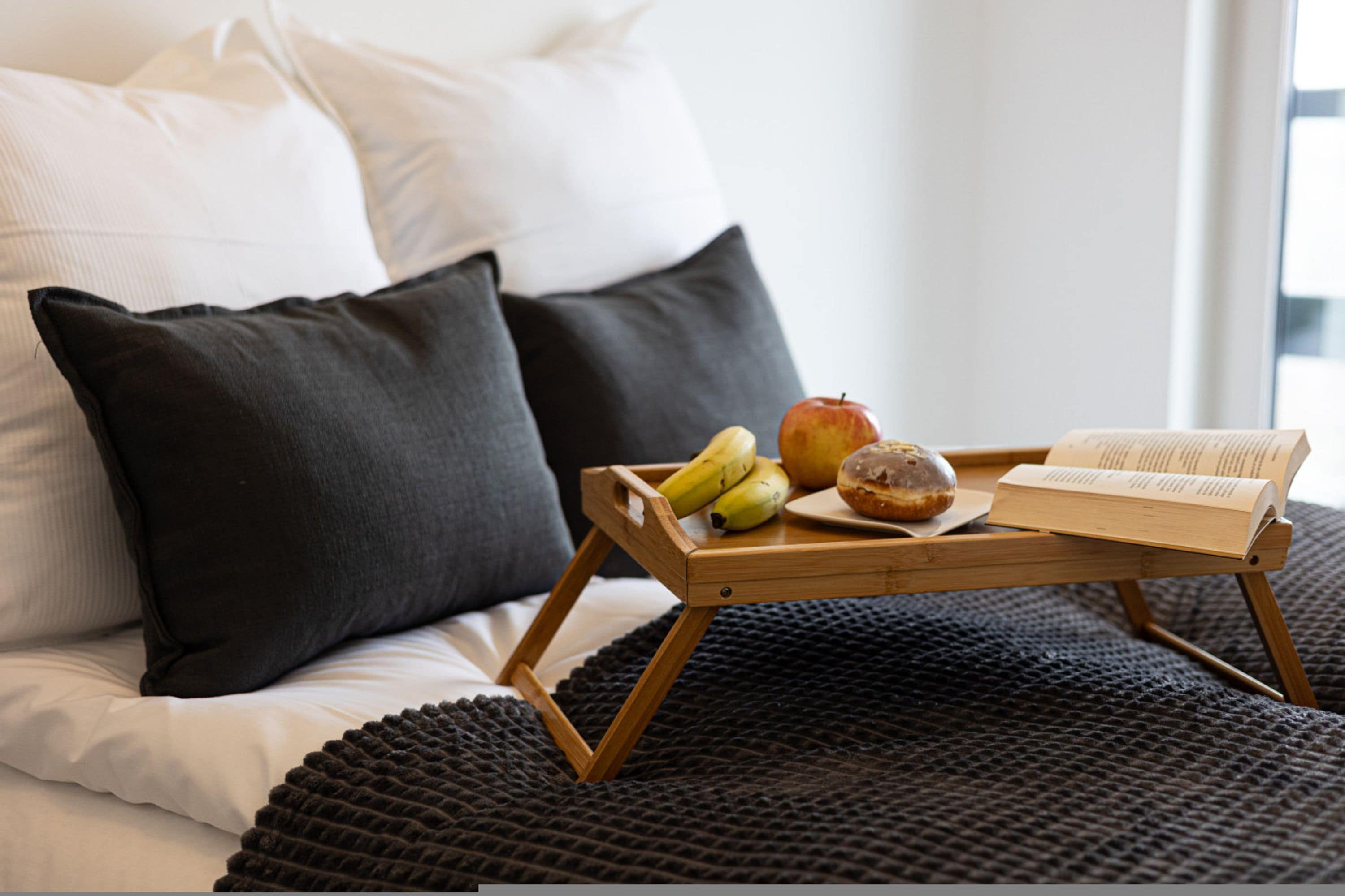 Cama com manta escura, bandeja de madeira com frutas e um livro, e almofadas.