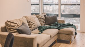 Un sofá seccional beige con varios cojines y una manta verde, ubicado en una habitación con piso de madera y ventanas amplias que ofrecen vista a un paisaje urbano.