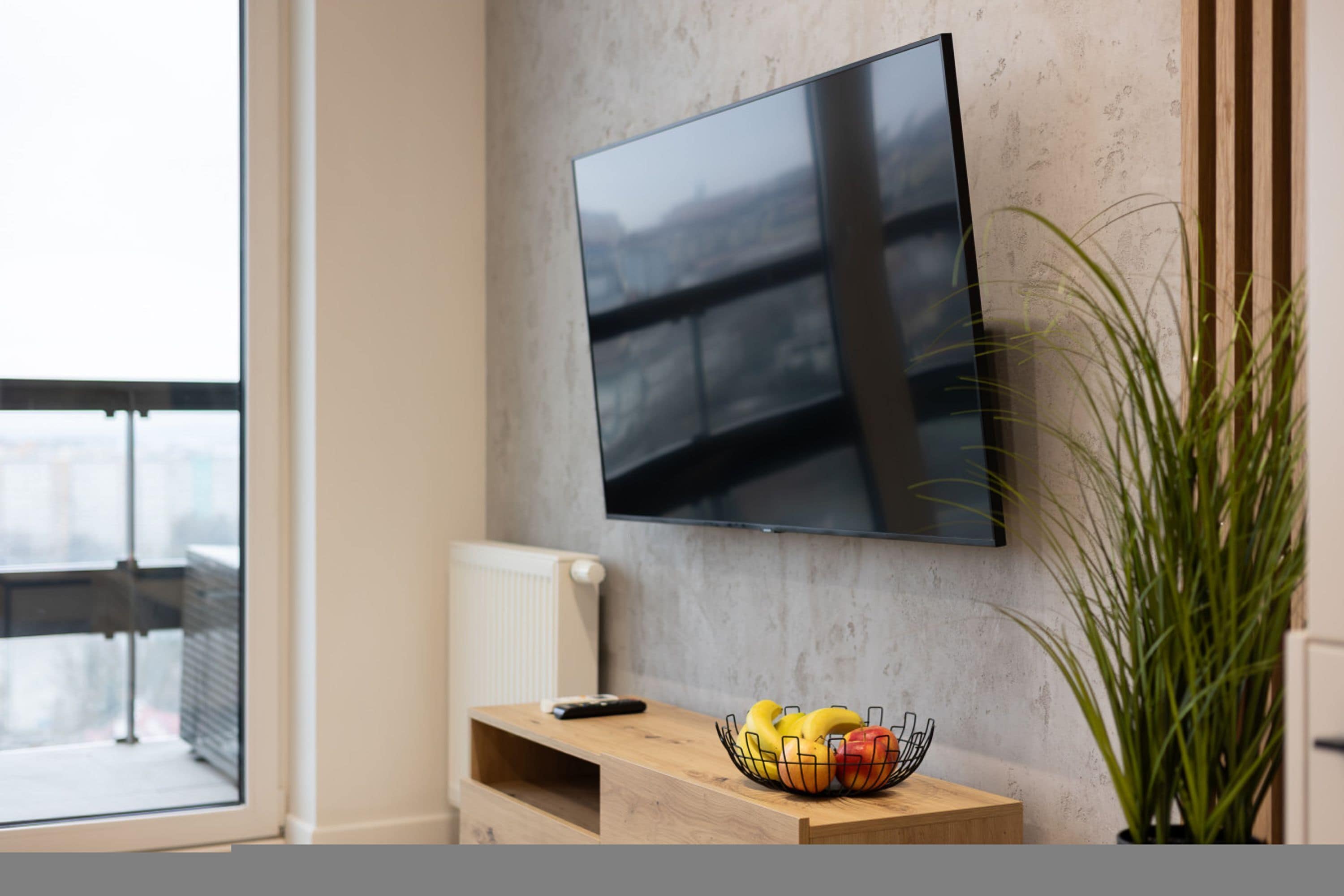 Un salon moderne avec un téléviseur à écran plat fixé au mur, une petite table en bois sur laquelle se trouve un panier de fruits et une plante en pot.