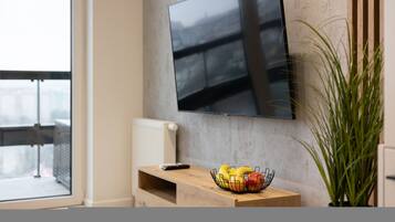 Un salon moderne avec un téléviseur à écran plat fixé au mur, une petite table en bois sur laquelle se trouve un panier de fruits et une plante en pot.