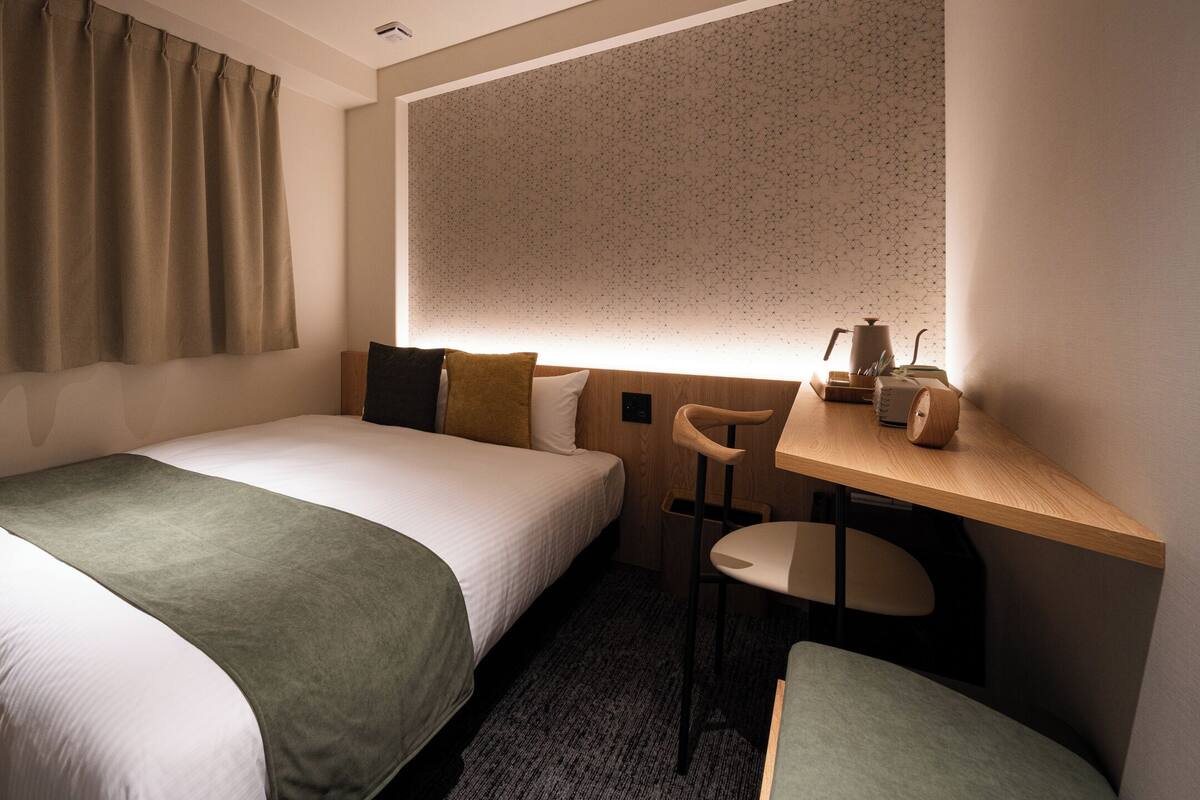 A hotel room with a single bed, a desk, a chair, and a small table with a lamp.