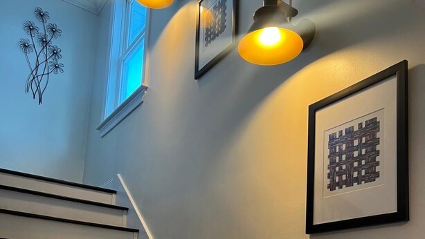 A staircase with white steps and dark handrails, flanked by wall-mounted light fixtures and framed artwork. - Adorable 4-bedroom house with AC in charming Attleboro (Attleboro)