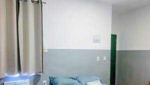 Een eenpersoonskamer met houten vloer, een bed met blauw beddegoed, een wandairconditioning en een raam met gordijnen.