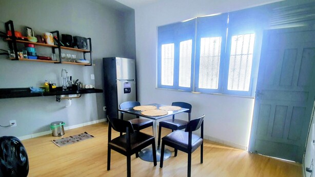 Een kleine keuken met een koelkast, een eettafel met stoelen en een raam met zonwering.