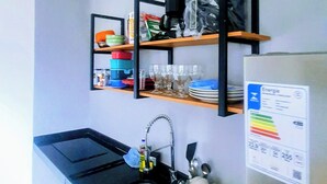 Een compacte keuken met een zwart aanrecht, een roestvrijstalen spoelbak en een koelkast. Daarboven bevinden zich planken met diverse keukenbenodigdheden en daaronder een kleine prullenbak.