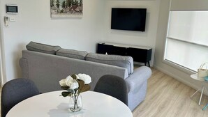 Ein modernes Wohnzimmer mit einem runden weißen Tisch, grauen Sofas, einem Flachbildfernseher und einem Gemälde mit einer tropischen Szene.