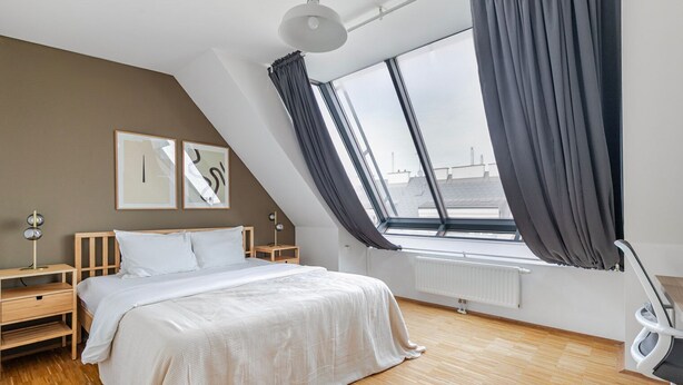Une chambre moderne avec un plafond en pente, une grande fenêtre avec des rideaux, un lit recouvert de linge de maison blanc, une table de chevet avec une lampe et des œuvres d’art accrochées au mur.