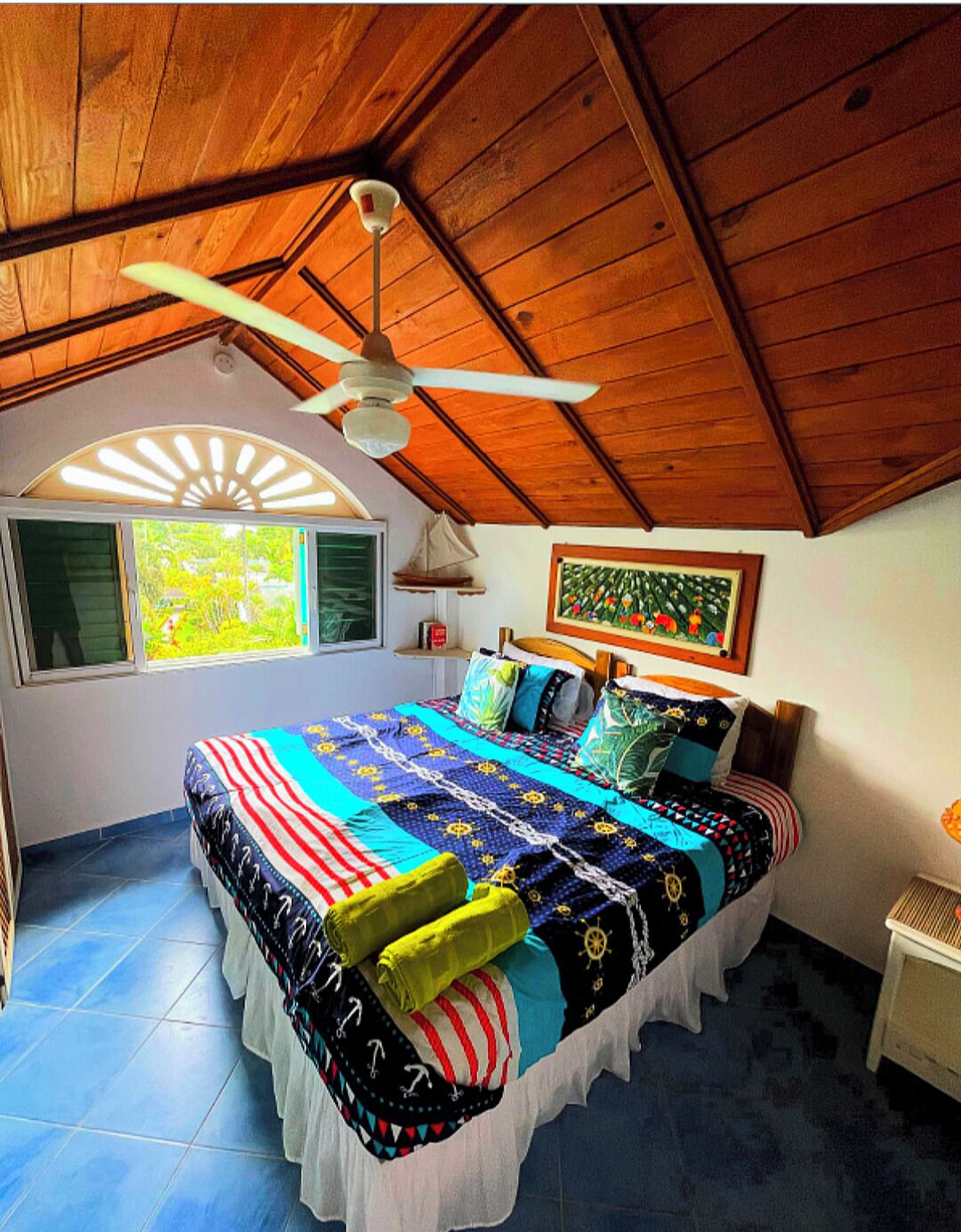 Un dormitorio con techo de madera, una cama con colcha colorida, un ventilador de techo y una ventana con vistas a la vegetación.