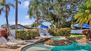 Un área de piscina con sillones de descanso, una chimenea de piedra y jardines tropicales.