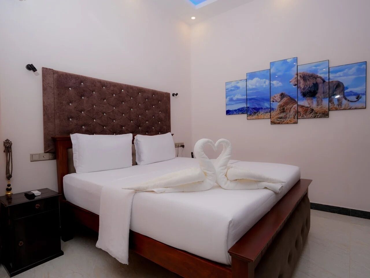 A neatly made bed with white linens and a heart-shaped towel arrangement, a wooden headboard, a bedside table with a lamp, and a wall with three framed pictures.
