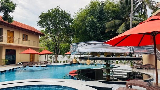 A swimming pool with lounge chairs and umbrellas, surrounded by trees and a building with a red roof. - Infenta's Villa (Pandatharippu)