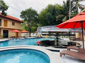 Une piscine entourée d’arbres, avec des chaises longues et des parasols, et un bâtiment doté d’un toit rouge.