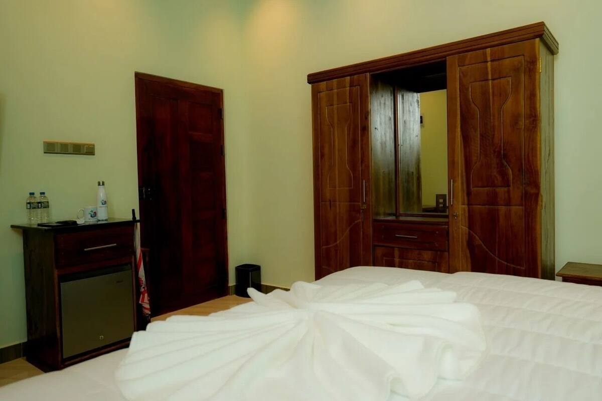A hotel room with a bed, a wooden wardrobe, a small desk with a mirror, and a wall-mounted light switch.