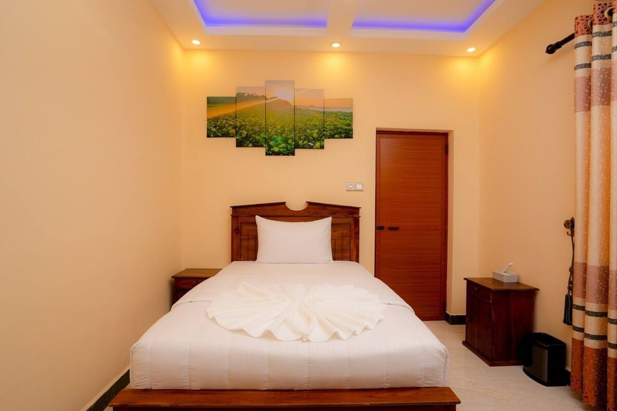A neatly made bed with a white comforter and a decorative towel arrangement, a wooden headboard, a wall-mounted painting of a landscape, a wooden door, and a small bedside table with a lamp.