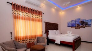 A hotel room with a bed, two armchairs, a round ottoman, and a large window with curtains. - Infenta's Villa (Pandatharippu)