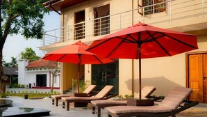 A poolside area with lounge chairs and red umbrellas, a building with a balcony, and a wooden door. - Infenta's Villa (Pandatharippu)