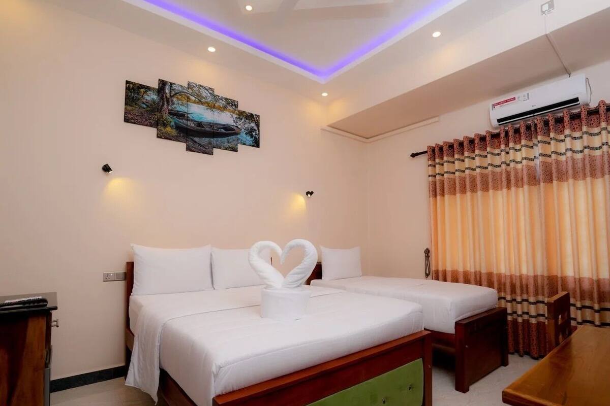 A hotel room with two beds, a green upholstered bench, a wooden table, an air conditioning unit, and a painting on the wall.