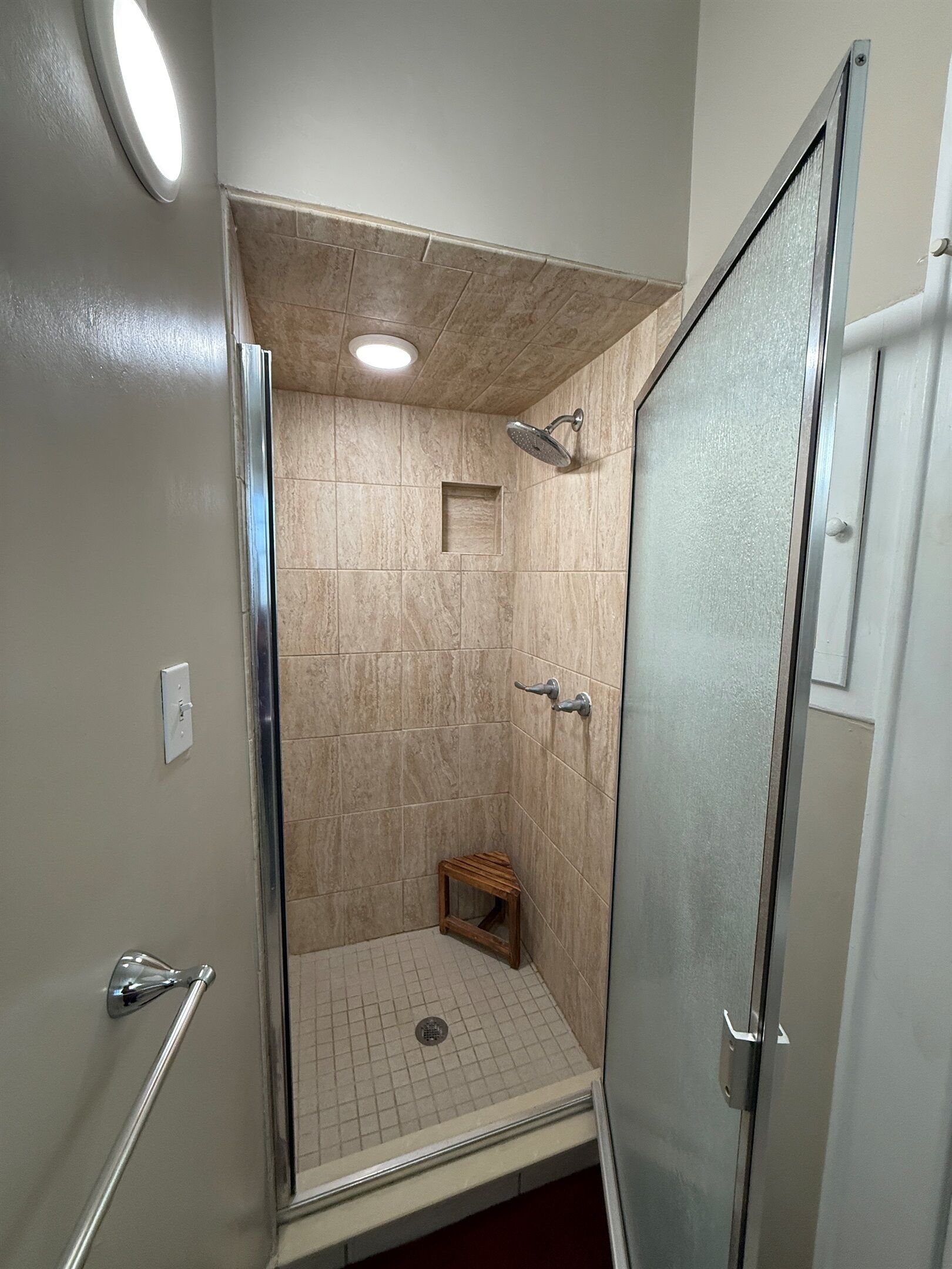 Une douche à l’italienne avec une porte en verre, un petit tabouret en bois et un sol carrelé.