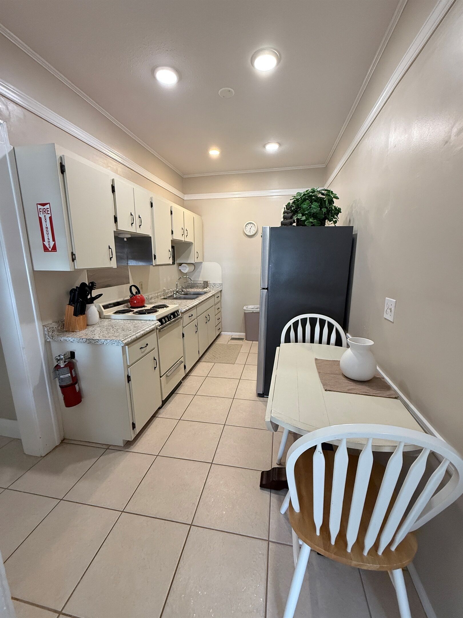 Une cuisine de taille réduite avec des meubles blancs, une cuisinière et un évier. On y trouve une petite table avec une chaise, un réfrigérateur et une plante en pot.