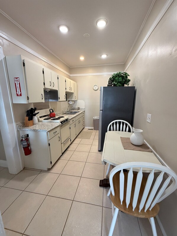 A compact kitchen with white cabinets, a stove, and a sink. There's a small table with a chair, a refrigerator, and a potted plant.