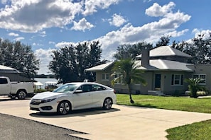 Un barrio residencial con un coche blanco estacionado en la entrada, una casa de techo gris y un cielo despejado.