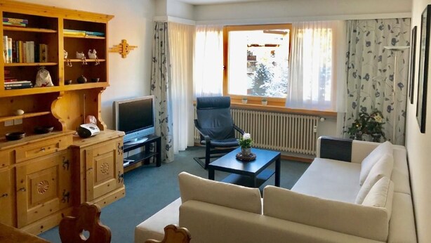 A living room with a sofa, a television, a wooden cabinet with books, and a window with curtains. - Derby 31 by Interhome (St. Moritz)