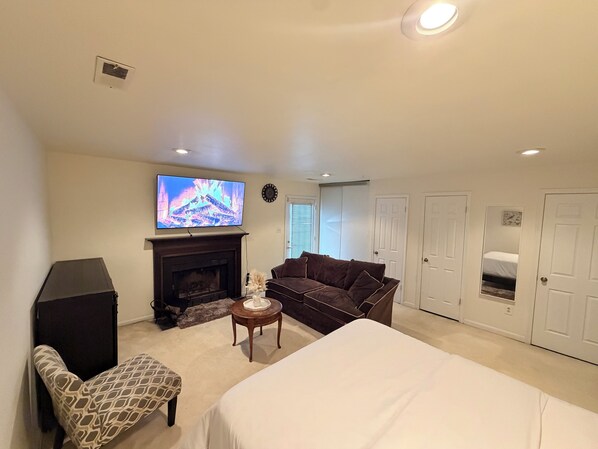 A spacious living room with a fireplace, a large flat-screen TV, a sofa, a patterned armchair, and a bed.