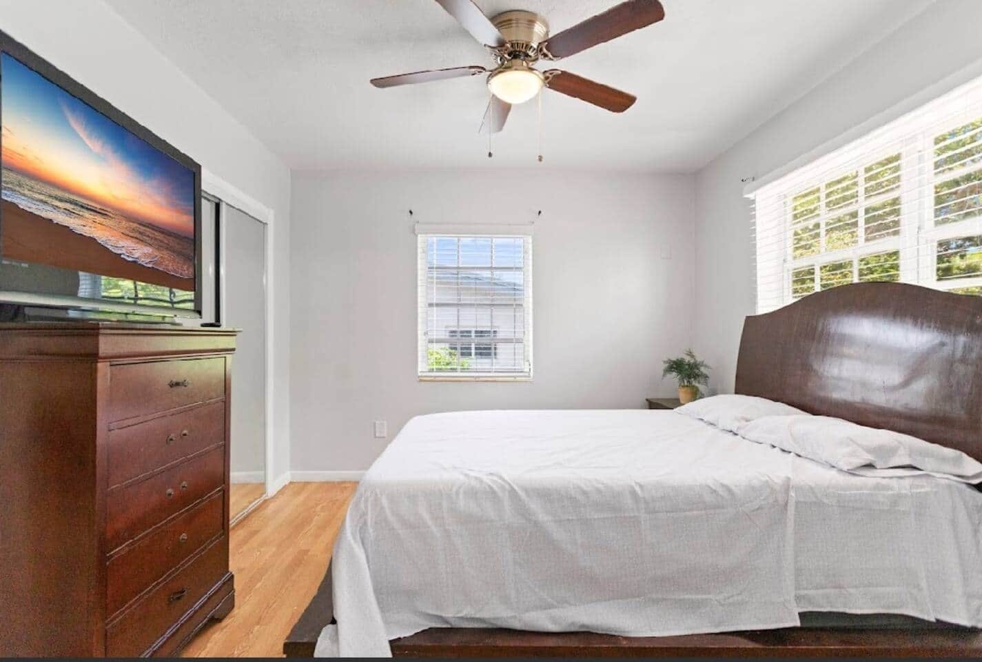 A bedroom with a bed, a ceiling fan, a dresser, a window, and a TV on the wall.