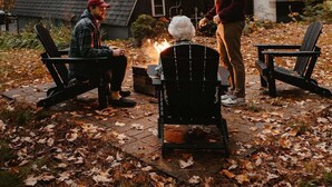 Hyggeligt udendørs område med Adirondack-stole, bålsted og en rustik hytte i baggrunden.