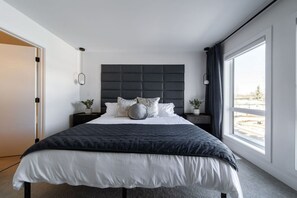 A modern bedroom with a large bed, a dark headboard, and a view of the outdoors through a large window. - Skyline Serenity | HotTub | Rooftop Patio |Central (Edmonton)