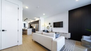 Una sala de estar moderna con un sofá, un televisor montado en la pared y una cocina al fondo.