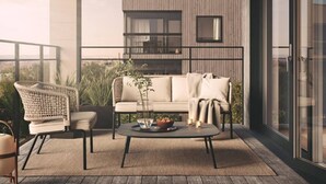 Et moderne udendørs opholdsområde med sofa, lænestol og sofabord, med udsigt til en bygning med store vinduer og en balkon.