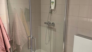 Et brusehækket brusebad med håndbruser og håndvask, et hvidt toilet og et håndklædestativ med lyserøde håndklæder.