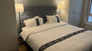 En pænt seng med hvide sengetøj og dekorative puder, en grå polstret sengegavl og en hvid garderobe.