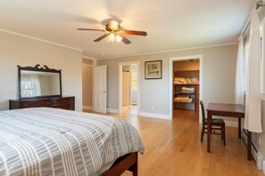 Ein Schlafzimmer mit Bett, Kommode, Schreibtisch und Stuhl. Ein Deckenventilator und eine Tür zu einem anderen Raum sind vorhanden.