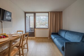 Ein Zimmer mit Esstisch, Stühlen, einem blauen Sofa, einem an der Wand montierten Fernseher und einem Fenster mit Vorhängen.