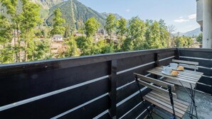 Un balcón con mesa y sillas, con vista a un paisaje montañoso con árboles y edificios.