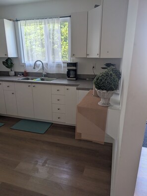 Een keuken met witte kastjes, een spoelbak, een koffiezetapparaat en een raam met dunne gordijnen.