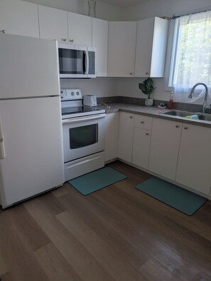 Une cuisine avec des meubles blancs, un réfrigérateur, une cuisinière, un four à micro-ondes, un évier et une fenêtre sur le plan de travail, où se trouve une plante.