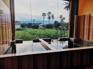 A modern bathroom with a glass-enclosed shower, wooden paneling, and a view of a lush green landscape with palm trees and houses. - Yogika-Center (Karangasem)