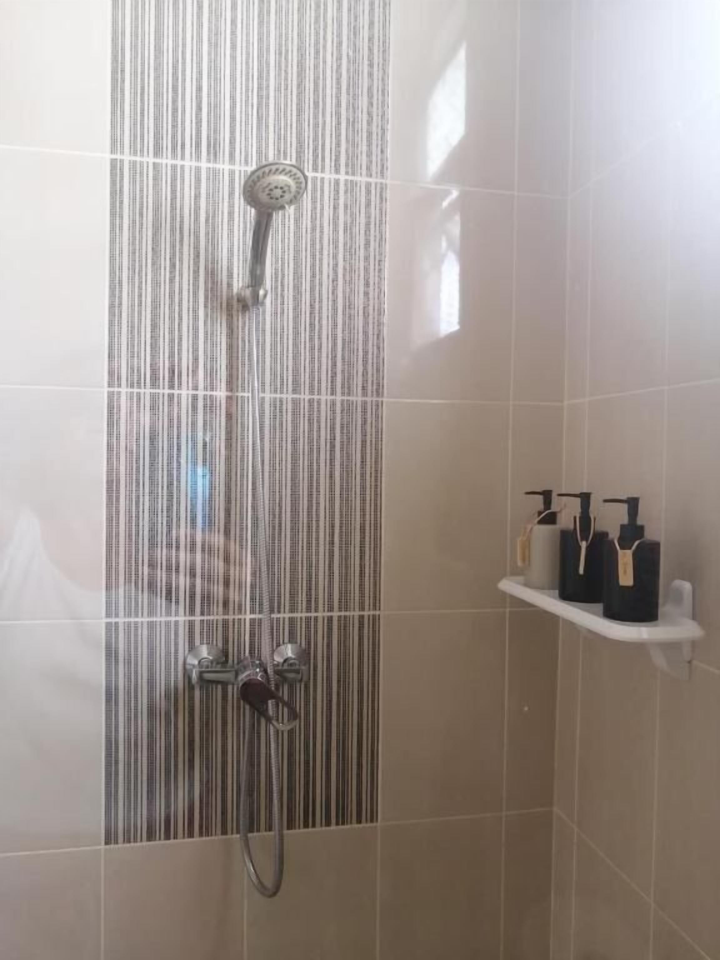 Una ducha con un panel de vidrio a rayas verticales, un cabezal de ducha de mano y un grifo instalado en la pared. Hay un estante con artículos de tocador.