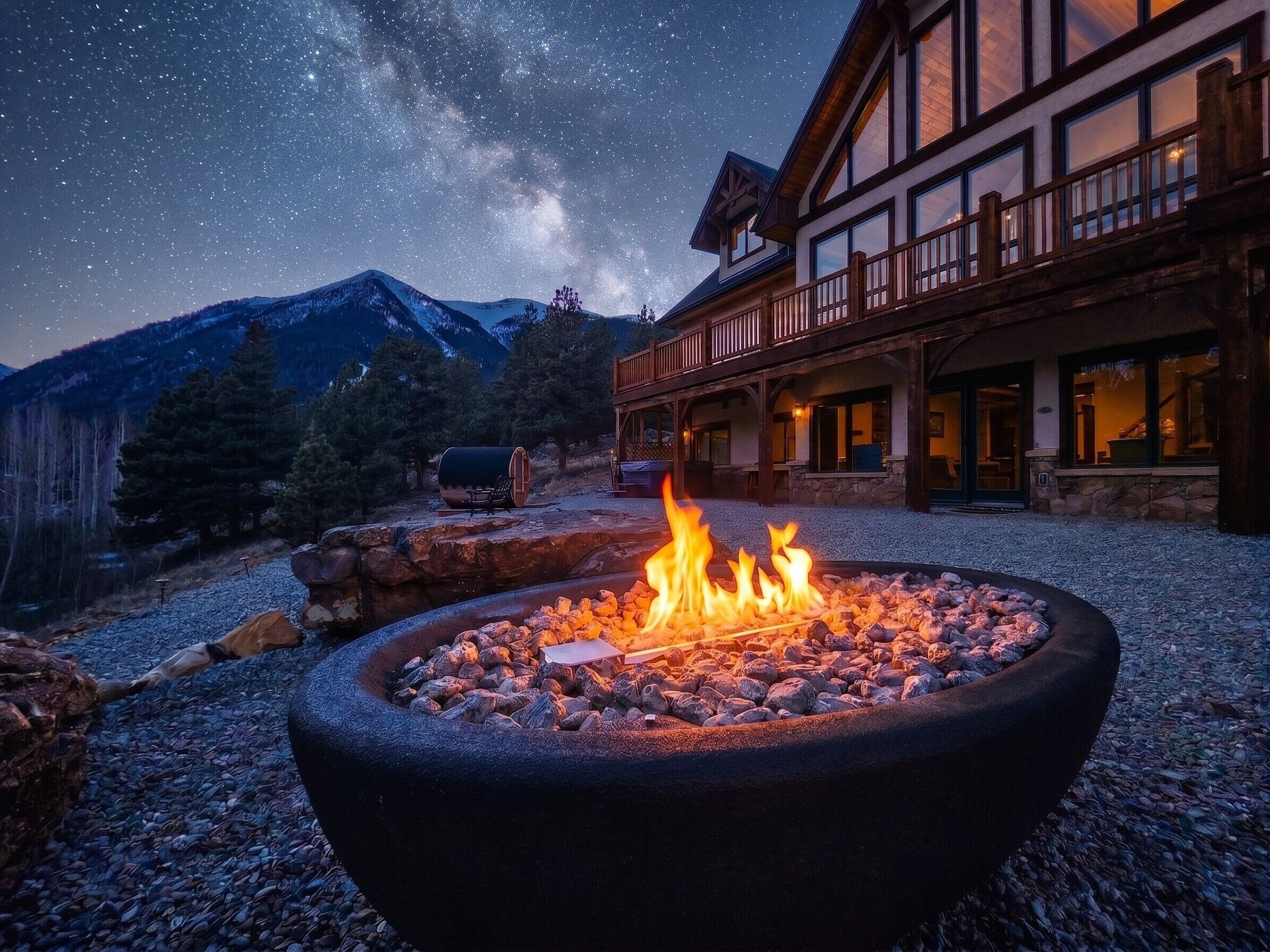 Un hotel con una fogata en primer plano, montañas al fondo y un cielo estrellado.