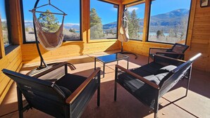 Eine Holzhütte mit großen Fenstern und Blick auf die Berge, einer Hängematte und zwei Liegestühlen.
