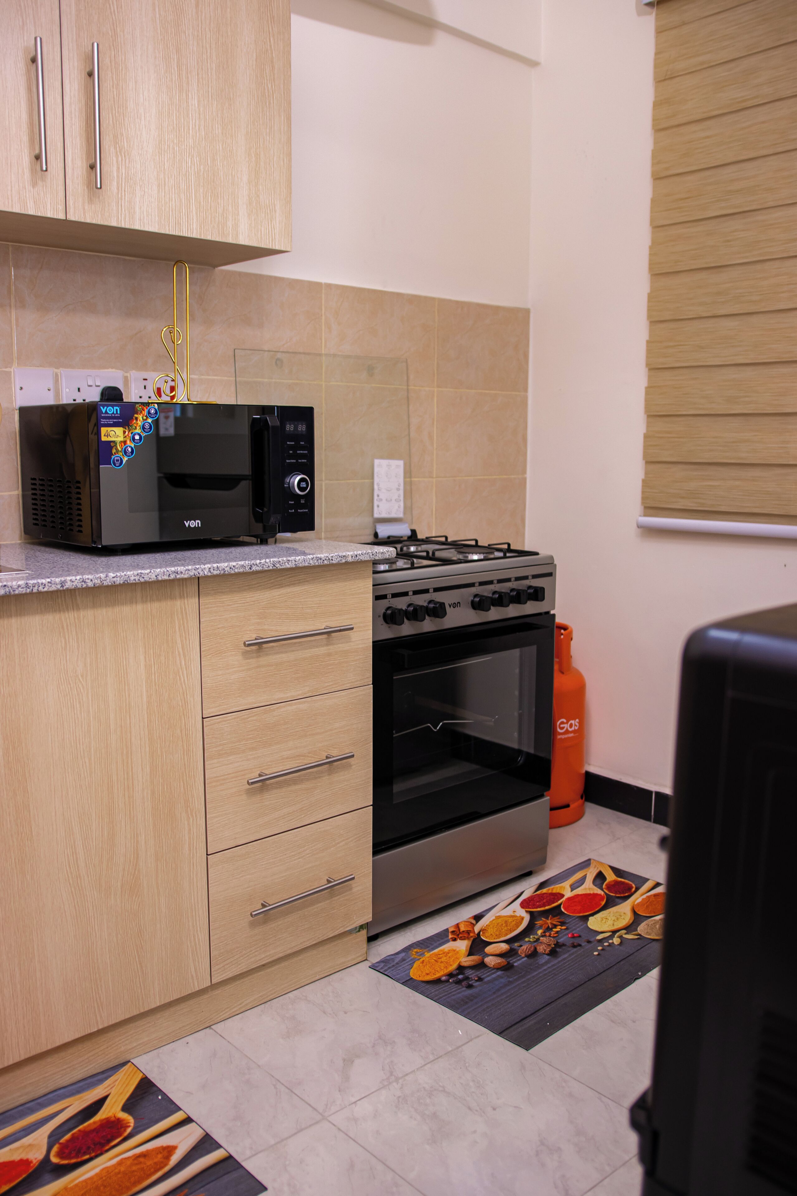 A kitchen with a microwave, stove, and gas stove. There are cabinets, a gas cylinder, and a rug with kitchen utensils.
