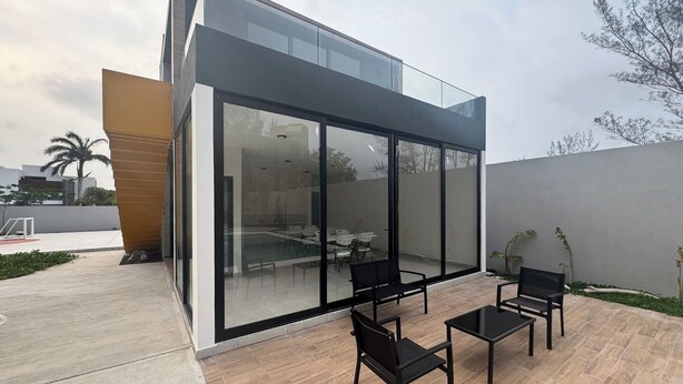 Um edifício moderno com fachada de vidro, área externa com assentos e mesas pretas, e piscina visível.
