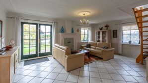 A spacious living room with a fireplace, two sofas, a coffee table, and a chandelier. There is a staircase to the right and a view of the outdoors through large windows. - Charming 5-person Villa by the sea (Callantsoog)