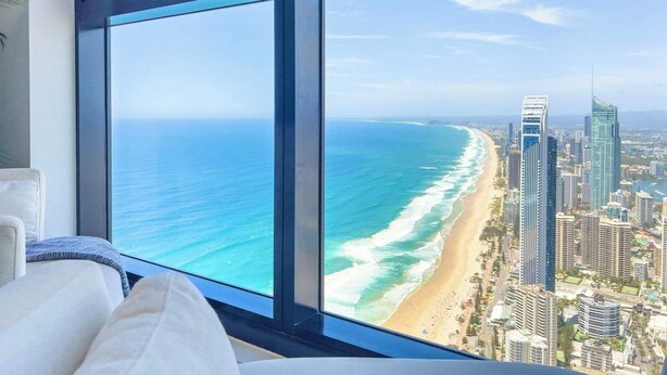 Habitación de hotel moderno en un edificio alto, con un amplio ventanal que ofrece vistas a la playa y al paisaje urbano.