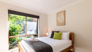 Ein Schlafzimmer mit einem großen Fenster, einem Bett mit grauer Decke, weißen und grünen Kissen und einer gerahmten Blattform-Kunst auf der Wand.