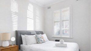 A bedroom with a bed, a bedside table with a lamp, and a window with blinds. - Luxury Spacious 3BR Family Home Walk To Harbour Bridge/ Opera House& CBD (Millers Point)