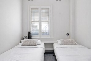 Une chambre avec deux lits simples, une fenêtre avec des stores, une table de chevet avec une lampe et un radiateur.